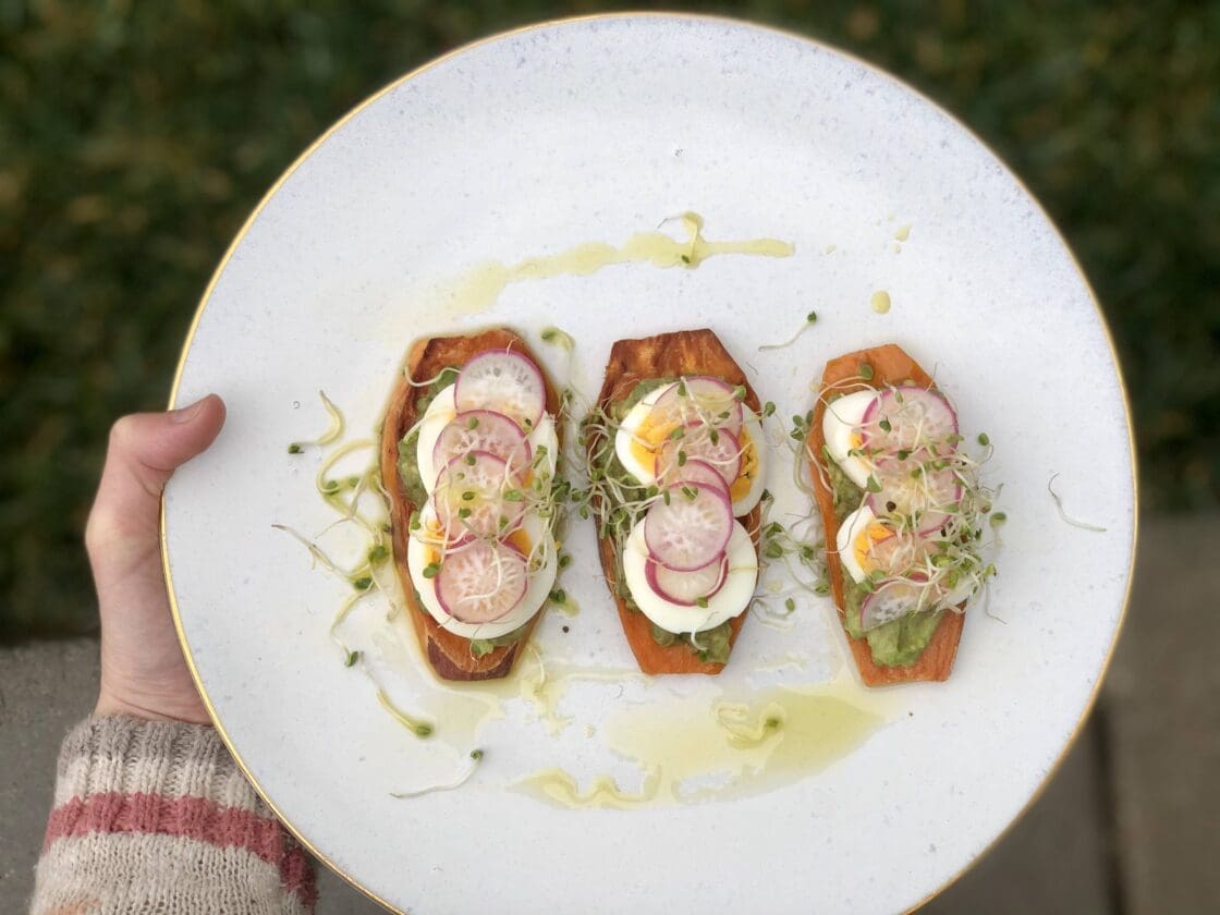 Hand holding plate of sweet potato toast with hard boiled egg, sprouts and avocado.