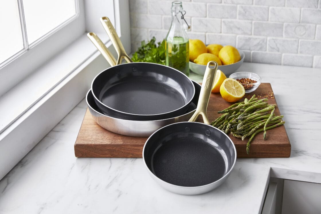 Image of the GP5 Stainless frypan 3-piece set with is shiny and stainless on the outside, with champagne handles and a nonstick cooking surface on a marble countertop with a marble subway backsplash. The pans sit atop a wood chopping block with asparagus and fresh lemon.