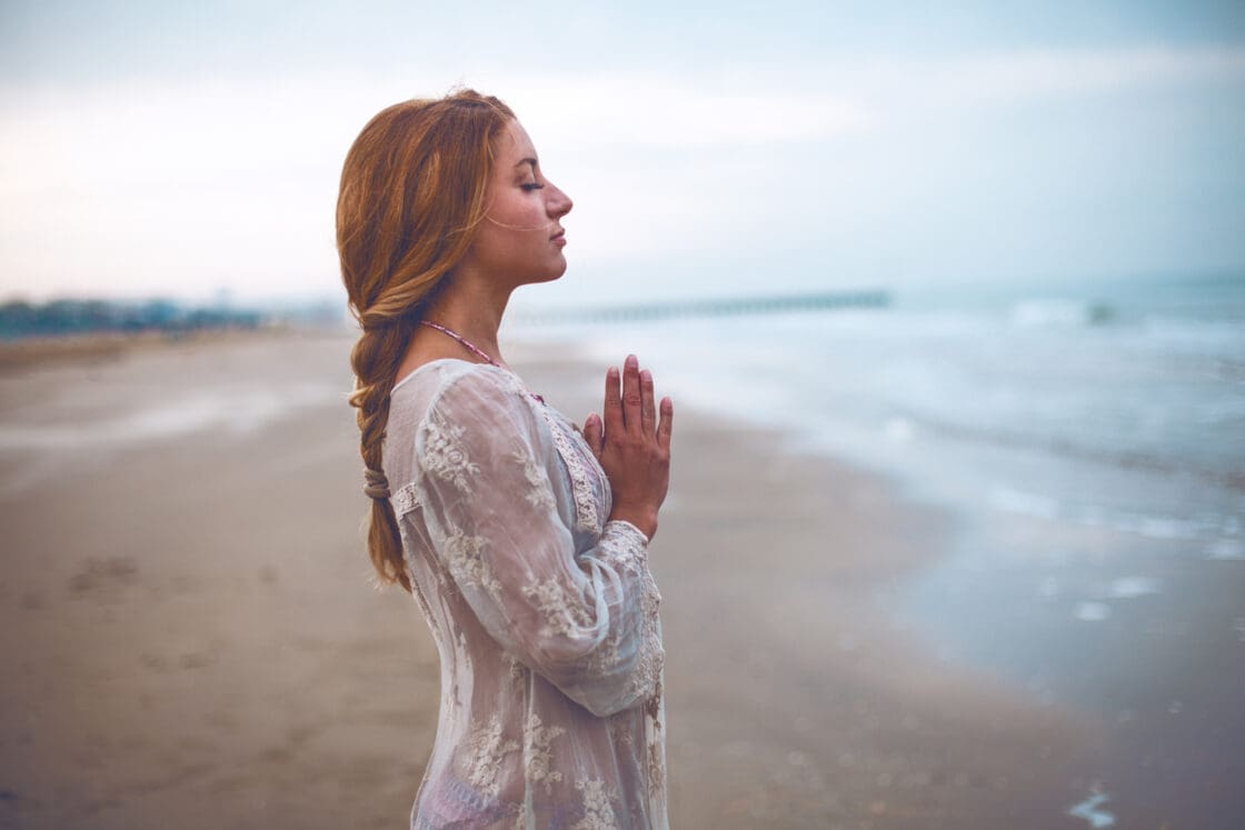 Image of woman standing in prayer pose on the beach, her eyes are closed doing Simple Breathwork Meditation For Beginners