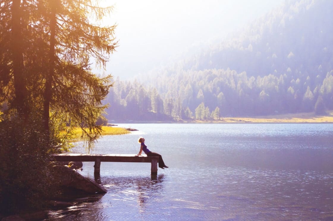 Image of a woman leaning back on her hands at the end of a pier on a lake with evergreens and a glowing light coming from the sky doing simple breathwork meditation.