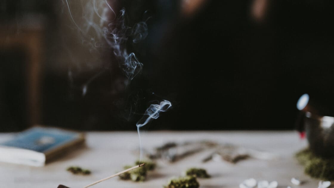 Image of incense burning safely in the foreground, with various wholistic, meditative items on the table behind with a journal showing that you can burn incense safely.