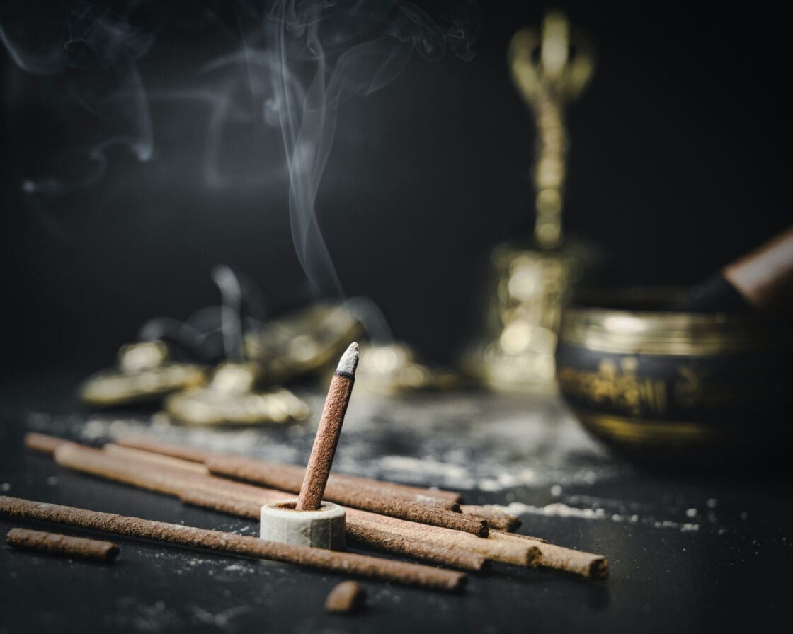 Image of incense burning safely with more incense sticks scattered on dark table with a singing bowl in the background showing that incense can be burned in a safe way.