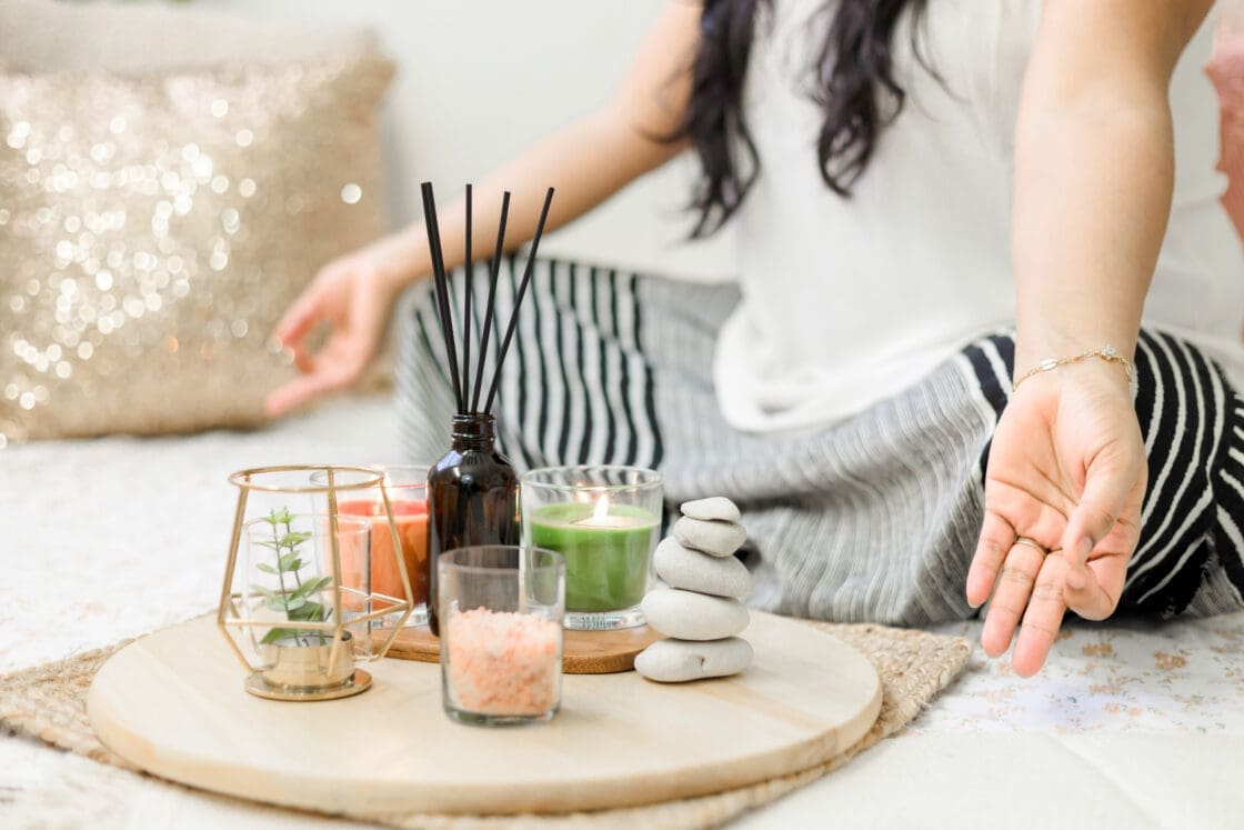 Image of woman meditating with legs crossed and hands in Gyana Mudra in front of her is a round wood plate that holds unlit incense, a stack of rocks and crystals.