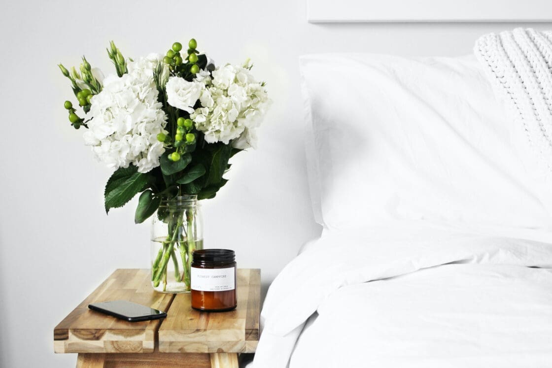 Image of White bouquet, candle, and phone next to bed with white sheets burning a candle instead of incense.