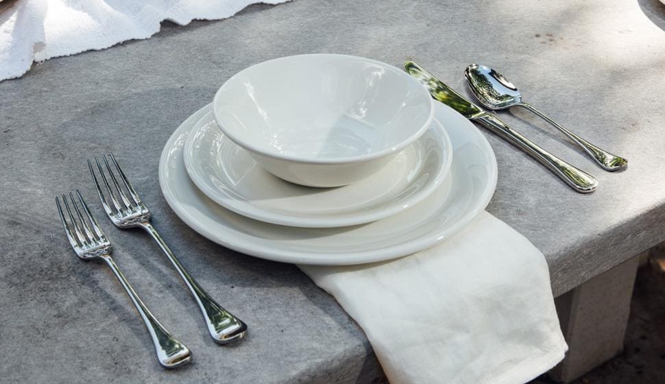 Image of non-toxic plateware by Made In, a white plate with a saucer stacked on it and a white bowl on that alongside shining silverware on a natural concrete table.