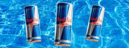 Image of 3 cans of Red Bull with a blue water background showcasing Red Bull ingredients but you'll want to know, are they safe to drink?