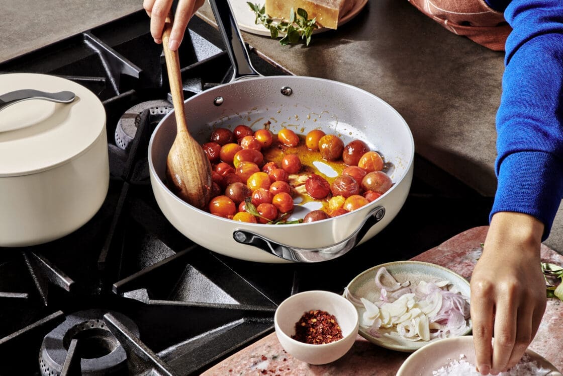 image of a Caraway set, Caraway home reviews on a stove with woman's hand reaching for salt. This cream set is different from the Caraway stainless steel sets.