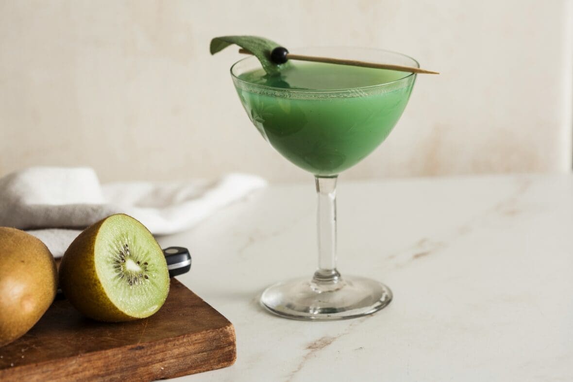 Image of a green cocktail in a coupe glass with garnish and a cut kiwi fruit on a cutting board.