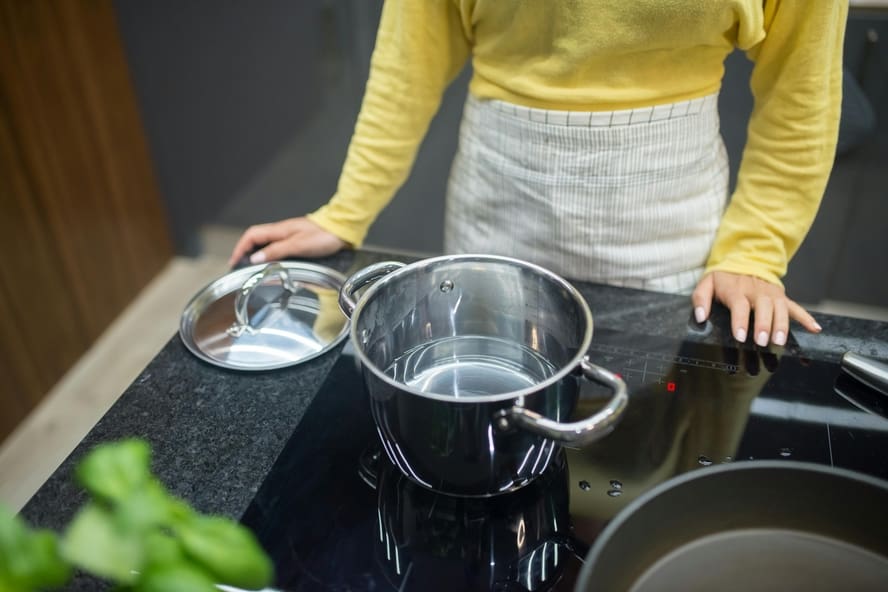 Image of a woman in a yellow sweater and apron leaning over an induction stovetop with a stainless steel pot and lid in front of her.