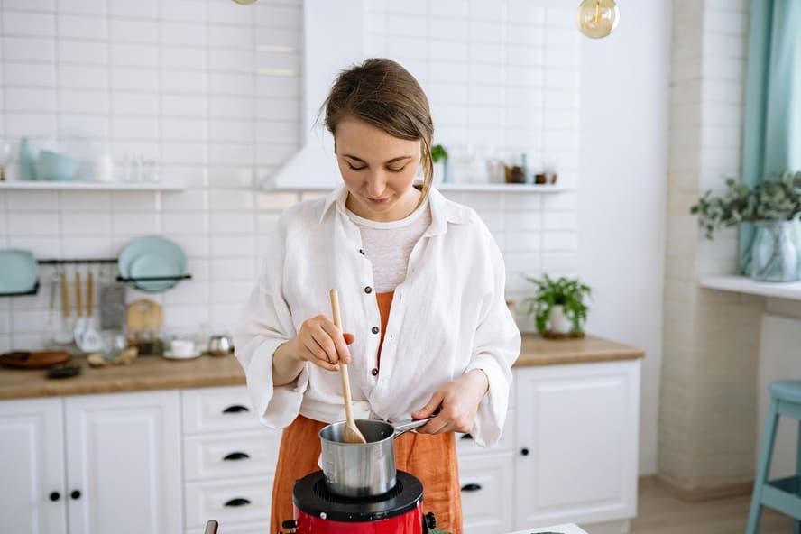 Image of a woman in a bright, minimalist kitchen using a wooden spoon to stir some food in a small stainless steel pot.