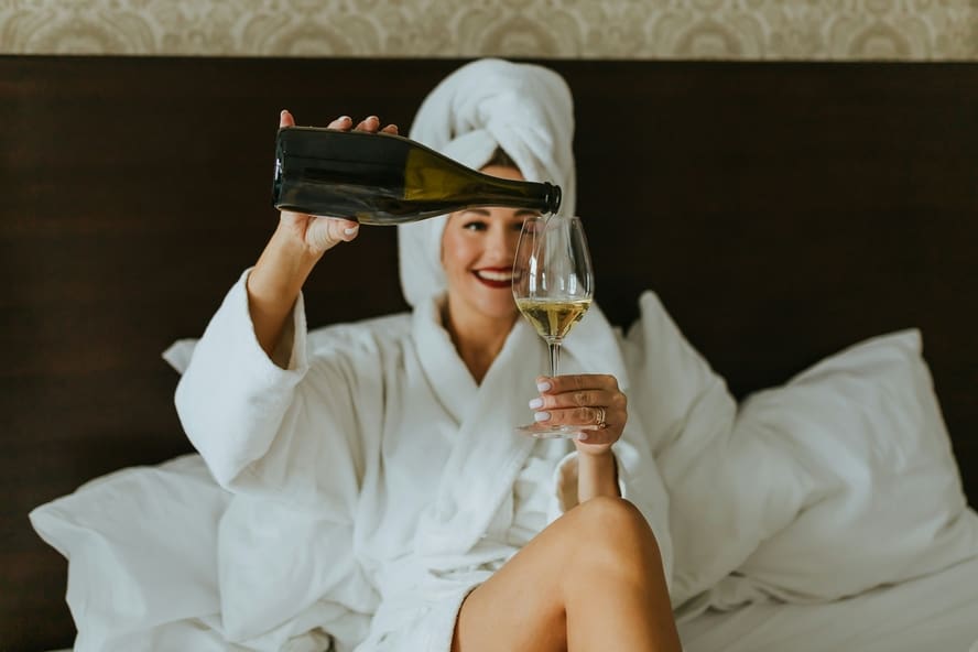 Image of a woman in a hotel room, wearing a white bathrobe and a towel around her head as she pours wine into a glass.