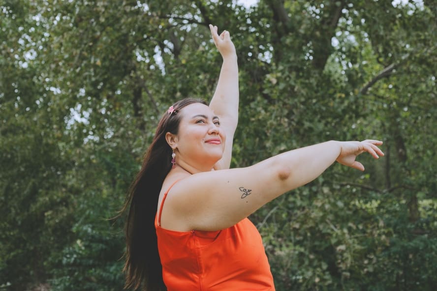 Image of a woman with long brown hair dancing in the park with her arms raised in an orange dress. Manifesting abundance can be done simply by being your true self.