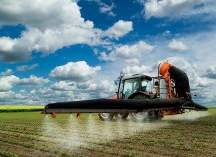Image of an agricultural machine in the fields under a blue sky with clouds, spraying the herbicide paraquat on crops.