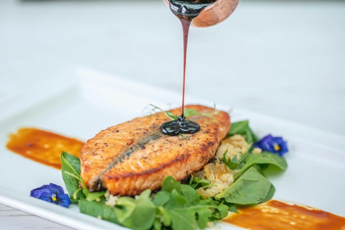 Image of a salmon filet served on a bed of spinach as someone pours a dark sauce over the dish.