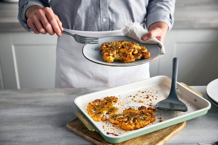 Image of a GreenPan Reserve baking sheet with a giant roasted cauliflower and someone serving it onto a plate.