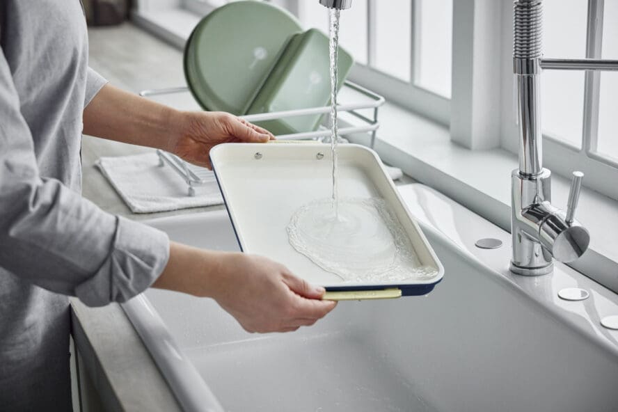 Image of someone holding a GreenPan cookie sheet by the gold handles and rinsing it under water in the sink.