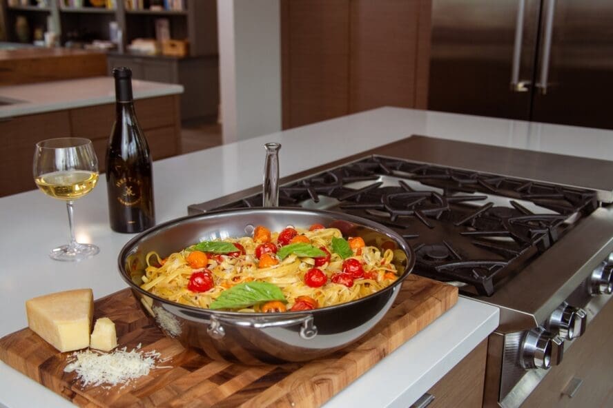 Image of a Hestan stainless steel Essential Pan on a wooden cutting board, filled with a tomato basil pasta and set alongside a block of cheese and a bottle of wine.