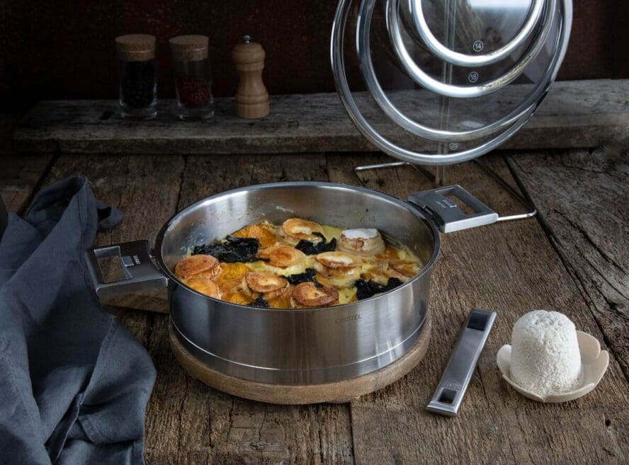 Image of a piece from the Cristel Strate 7-piece set, with the glass lids in the background and the detachable handle beside it.