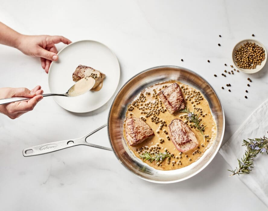 Image of a Demeyere Industry 5 frypan being used to cook steaks in a peppercorn sauce, with a pair of hands plating steak beside it.