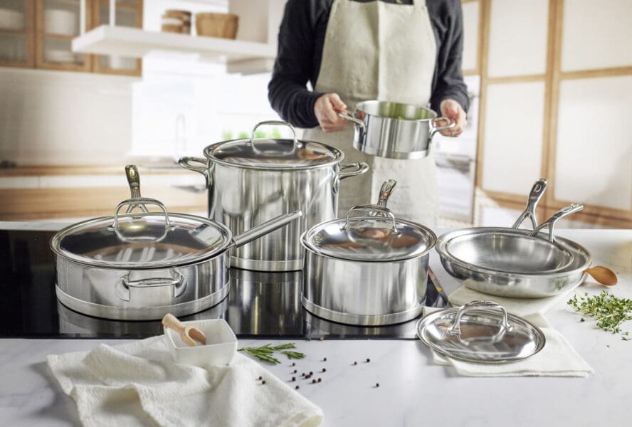 Image of several pieces in a Demeyere Atlantis set shown on a glass cooktop, with a home cook holding a stockpot behind the counter.