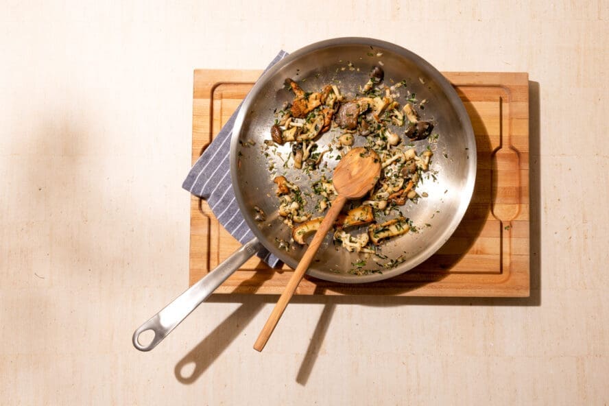 Image of a Made In stainless steel frying pan on a wooden cutting board with cooked mushrooms and a wooden spoon.