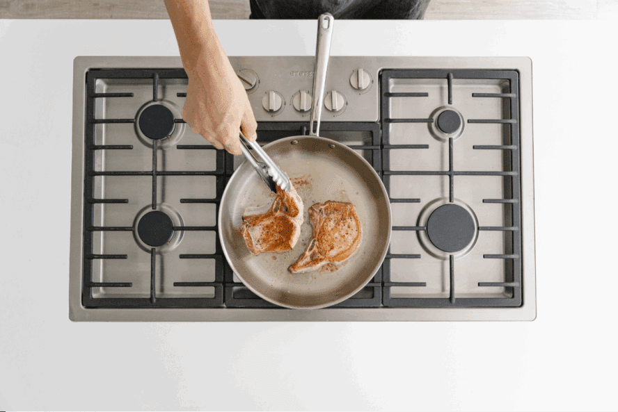 An overhead view of a Misen stainless steel skillet on a five-burner cooktop, with a pair of hands using tongs to sear pork chops.