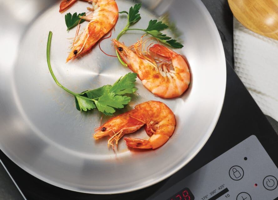 Image of three shrimp being pan-fried in a Tramontina stainless steel frypan on a glass cooktop.