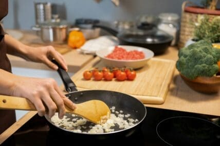 Image of someone cooking onions with a traditional nonstick pan on a glass stovetop, with tomatoes on a cutting board next to them.